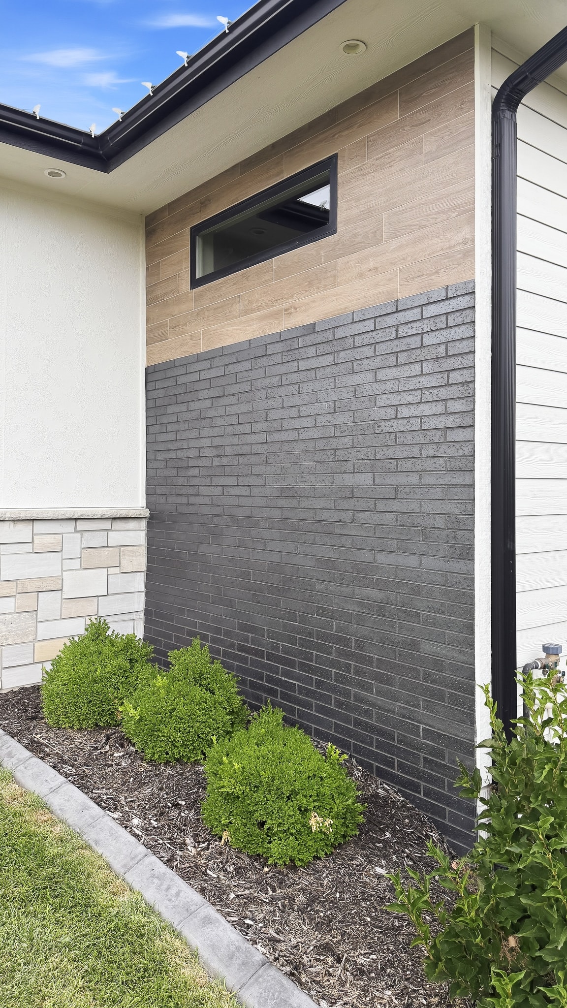 Modern house exterior corner with black brick-look tile, wood-grain tile panels and shrub landscaping
