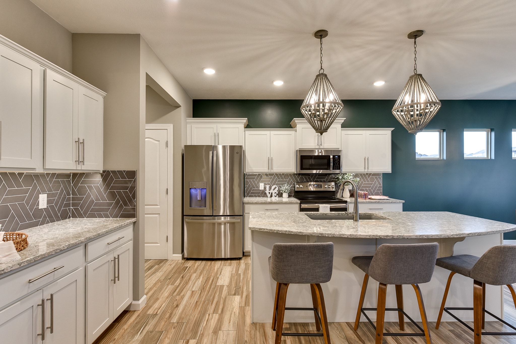 Kitchen with white cabinetry, teal accent wall, geometric gray tile backsplash, granite island and bead chandelier pendants