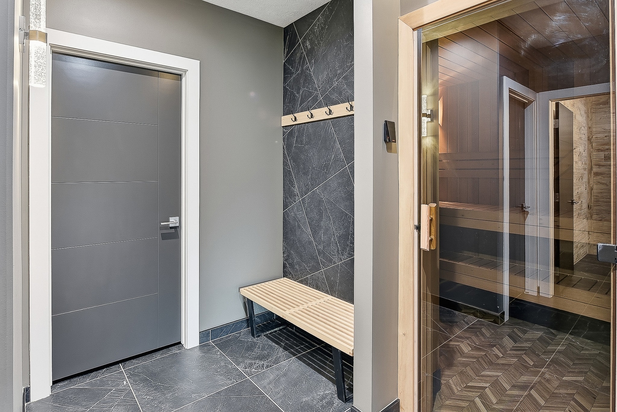 Sauna room entry with dark marble tile walls, wood bench, coat hooks and glass sauna door